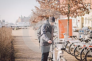 Man rents electric white bicycle in Copenhagen. E-Bicycle Rental Station. Male parked city bikes for rent, saying Save CO2 and