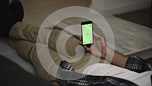 Man resting on a couch at home, using smartphone with green mock-up screen