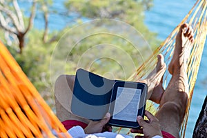 Man resting in hammock on seashore and reading ebook