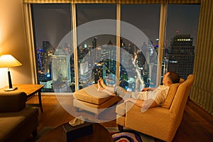 Man resting in room interior in the evening and looks through window on night city