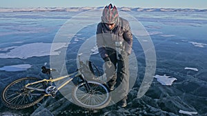 The man is resting sitting on the wheel of a bicycle on ice. He drinks tea from a thermos bottle. The cyclist is dressed