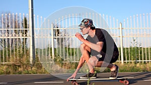 Man rides longboard sitting at empty straight road and touch his sunglasses. Summer vacation hipster rides skateboard