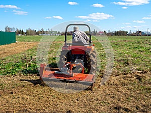 A man rides a tractor with a mower in a field. Mulching and mowing the grass. Clear day, blue sky. Land cultivation