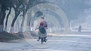 Man riding bicycle and carrying pillion whom he taking to school in winter foggy weather in village of Bangladesh