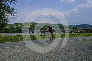 A man riding a bike on the bike lane across the mountains view, river and a bridge over the river. Langhirano, Italy