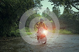 man riding small endurom motorcycle crossing shallow creek among rain falling at forest