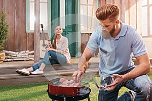 Man roasting meat on barbecue grill with woman with wine behind