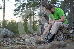 Man roasts a sausage over a fire in a metal camp stove and holds a mug of hot drink in hands. Outdoor cooking.