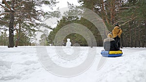 Man is rolling woman on tubing by snow in winter park, back view.