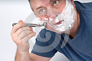 A man's face in shaving foam shaves with a safety razor