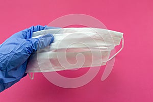 A man`s hand in a blue medical glove holds a medical mask on a pink background. Selective focus