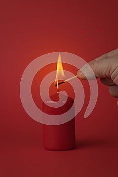 A man`s hand with a burning match lights a red candle on a red background