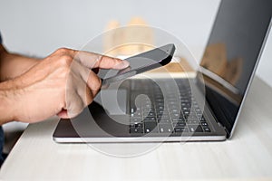 Man`s hand grasps smartphone and touches a screen with his fingers to remote power off a laptop. Working at home concept