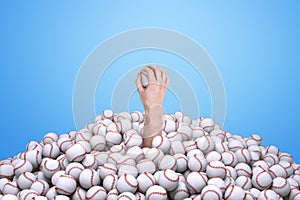 Man`s hand holding a baseball, emerging from under a big pile of baseballs.