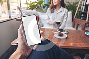 A man`s hand holding black mobile phone with blank white screen with woman sitting in cafe