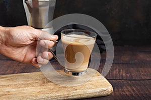 Man& x27;s hand holding cup of coffee with milk on wooden table and Italian coffee pot in the background