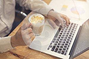 man's hand holding cup of hot latte coffee working with laptop c