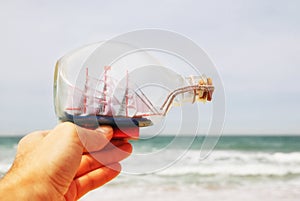 Man's hand holding decorative boat in the bottle in front of sea horizon. vintage filtered.