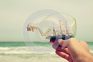 Man's hand holding decorative boat in the bottle in front of sea horizon. vintage filtered.