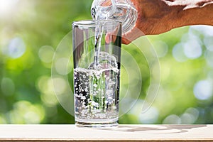 Man`s hand holding drinking water bottle water and pouring water into glass on wooden tabletop on blurred green bokeh background