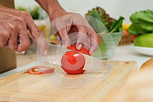 Man's hand is holding knife to thinly cut round, red tomato on a cutting board. To cook in the kitchen during the holidays