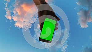 Man`s hand holding mobile smartphone with green screen on cloudy sky background and rotates