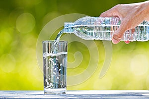 Man`s hand holding plastic bottle water and pouring water into glass on wooden table on blurred green bokeh background