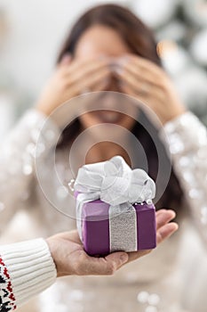 Man's hand is holding purple Xmas present with a ribbon in front of a woman closing her eyes with hands