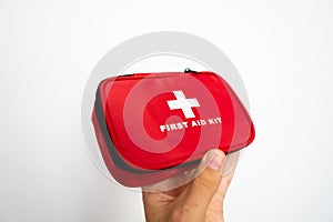 Man`s hand holding a red first-aid kit with a white wall on background.