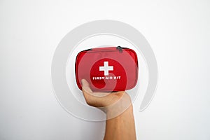 Man`s hand holding a red first-aid kit with a white wall on background.