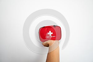 Man`s hand holding a red first-aid kit with a white wall on background.