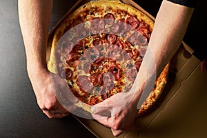 Man's hand is holding slice of double cheese pizza from whole Pizza in a cardboard box dark table background