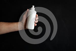 A man`s hand holding a white soap bottle on a black background. Unbranded blank bottle with liquid soap