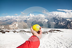 A man`s hand holds an apple against the background of snow-capped mountains and snow underfoot.