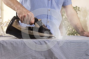 A man`s hand holds a black iron and irons the shirt on an ironing board. Household duties performed by men.