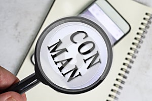 A man's hand holds a black magnifying glass by the handle with con man text.