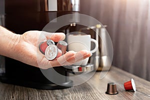 A man's hand holds capsules for a coffee machine against the background of a kitchen with cups and a coffee machine