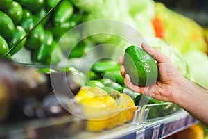 man& x27;s hand holds a fresh avocado next to a shelf with vegetables in a supermarket.