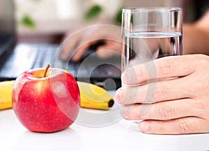 A man\'s hand holds a glass of water and an apple and a banana nearby. Healthy snack at work. Close-up. Selective focus