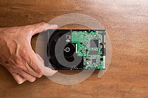 A man`s hand holds a hard disk memory located on a wooden base. The hard drive shows electronics and a printed circuit board