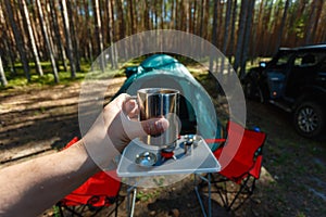 The man's hand holds an iron mug with a warm drink on the background of the table and chairs, campfire, a tent and an SUV in camp