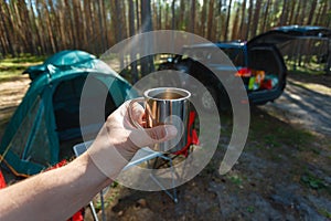 The man's hand holds an iron mug with a warm drink on the background of the table and chairs, campfire, a tent and an SUV in camp