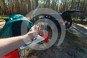 The man's hand holds an iron mug with a warm drink on the background of the table and chairs, campfire, a tent and an SUV in camp