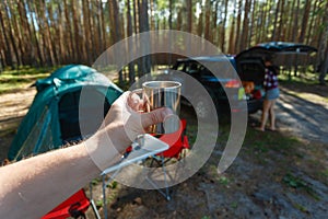 The man's hand holds an iron mug with a warm drink on the background of the table and chairs, campfire, a tent and an SUV in camp