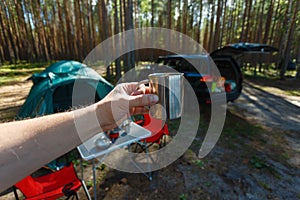 The man's hand holds an iron mug with a warm drink on the background of the table and chairs, campfire, a tent and an SUV in camp