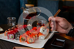 A man`s hand holds Japanese wooden sticks against the background of a sushi set and a glass teapot with tea in a cafe