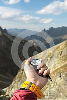 A man`s hand holds a pocket magnetic compass for navigation against the backdrop of a rocky slope and a mountain. The