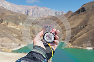 A man`s hand holds a pocket magnetic compass for navigation against the backdrop of a rocky slope and a mountain lake