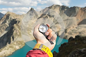 A man`s hand holds a pocket magnetic compass for navigation against the backdrop of a rocky slope and a mountain lake