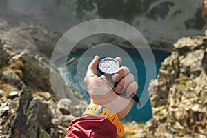 A man`s hand holds a pocket magnetic compass for navigation against the backdrop of a rocky slope and a mountain lake